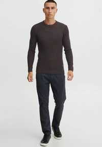 Dark brown ribbed long-sleeve sweater with a round neckline, paired with navy blue pants and black sneakers. Subtle brand logo detail visible.