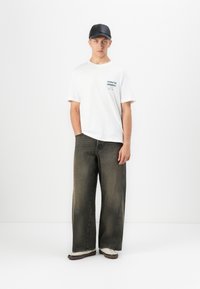 Ung mann iført svart caps, hvit t-skjorte med tekst, vide falmede svarte jeans og beige joggesko foran en ensfarget hvit bakgrunn.