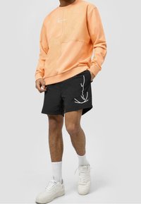 Sudadera naranja con un acabado texturizado y un logo blanco, combinada con pantalones cortos negros con arte lineal blanco, llevada con zapatillas y calcetines blancos.