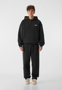 Crna dukserica s kapuljačom i odgovarajuće trenirke od mekanog materijala, s kengurU džepom i minimalnim logotipom na prsima. Cipele su bež.