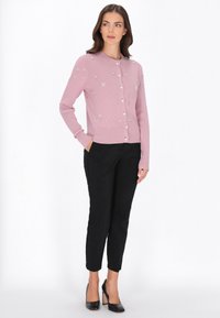 Világos rózsaszín, bordázott kardigán virágmintás díszítésekkel és gombokkal, fekete, testhezálló nadrággal és fekete magassarkú cipővel párosítva.