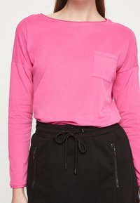 Rosa Langarm-T-Shirt mit weitem Ausschnitt und aufgesetzter Brusttasche, kombiniert mit einem schwarzen Rock mit Zugband und seitlichen Reißverschlusstaschen.