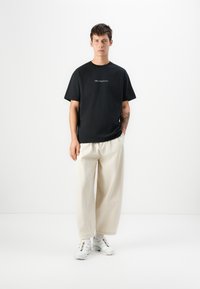 Svart bomulls T-skjorte med korte ermer og en subtil logo på brystet, kombinert med kremfargede vide bukser og hvite sneakers.