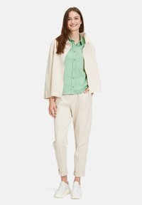 Cappotto crema sopra una camicia botton up verde chiaro, abbinato a pantaloni beige chiaro e sneakers bianche. Texture morbide in stile casual.