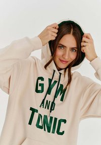 Béžová mikina vyrobená z bavlněné směsi, s potiskem "GYM AND TONIC" v tmavě zelené barvě. Má kapuci na šňůrku s kontrastním zeleným vnitřkem.