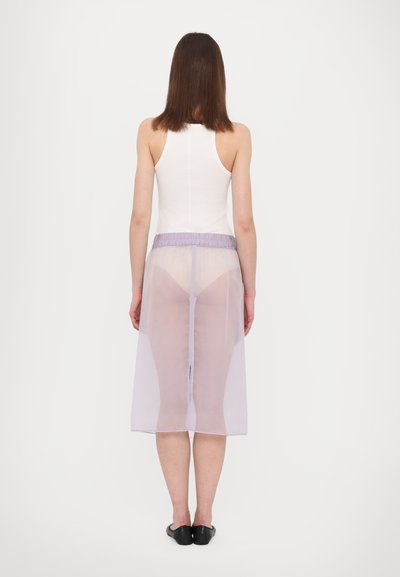Jupe violette transparente avec une taille élastique, design en couches et fente sur le côté, portée par-dessus un débardeur blanc et des chaussures noires.