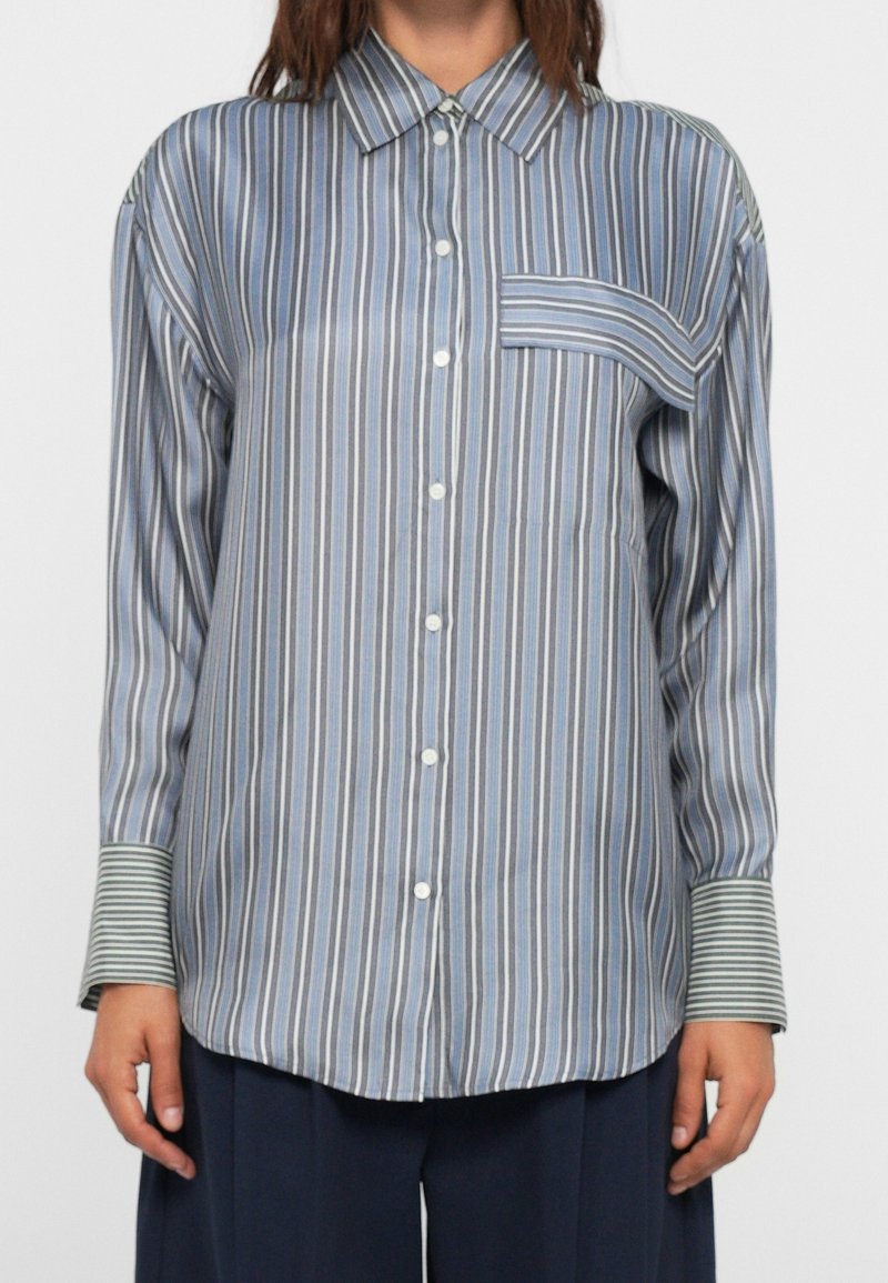 Femme portant une chemise ample à rayures bleues avec des poignets rayés contrastants et une poche poitrine horizontale, associée à un pantalon foncé.