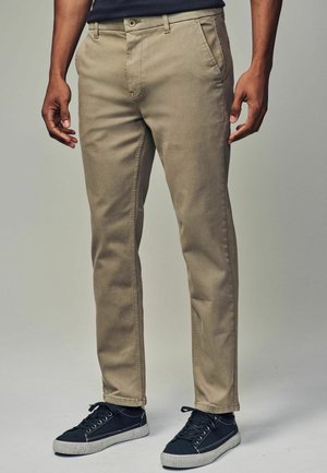 Beige Baumwoll-Chino-Hosen mit geradem Schnitt, ausgestattet mit Eingrifftaschen, Gürtelschlaufen und einem Reißverschluss. Getragen mit schwarzen Sneakers.
