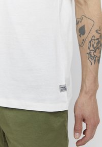 Hvit bomull t-skjorte med korte ermer, med en sidelabeldetalj og lett tekstur, kombinert med olivengrønne shorts, som viser en tatovert arm.