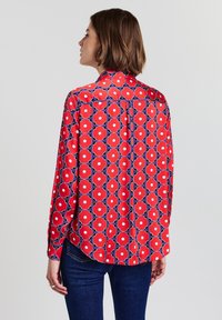 Rote Bluse mit langen Ärmeln und floralem Muster, weißen Mittelpunkten und blauen Konturen; verfügt über eine Knopfleiste am Rücken und einen lockeren Schnitt.
