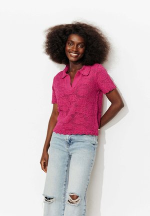 Jeune femme aux cheveux bouclés volumineux sourit, portant un haut rose vif texturé à manches courtes et un jean bleu clair déchiré sur fond blanc.