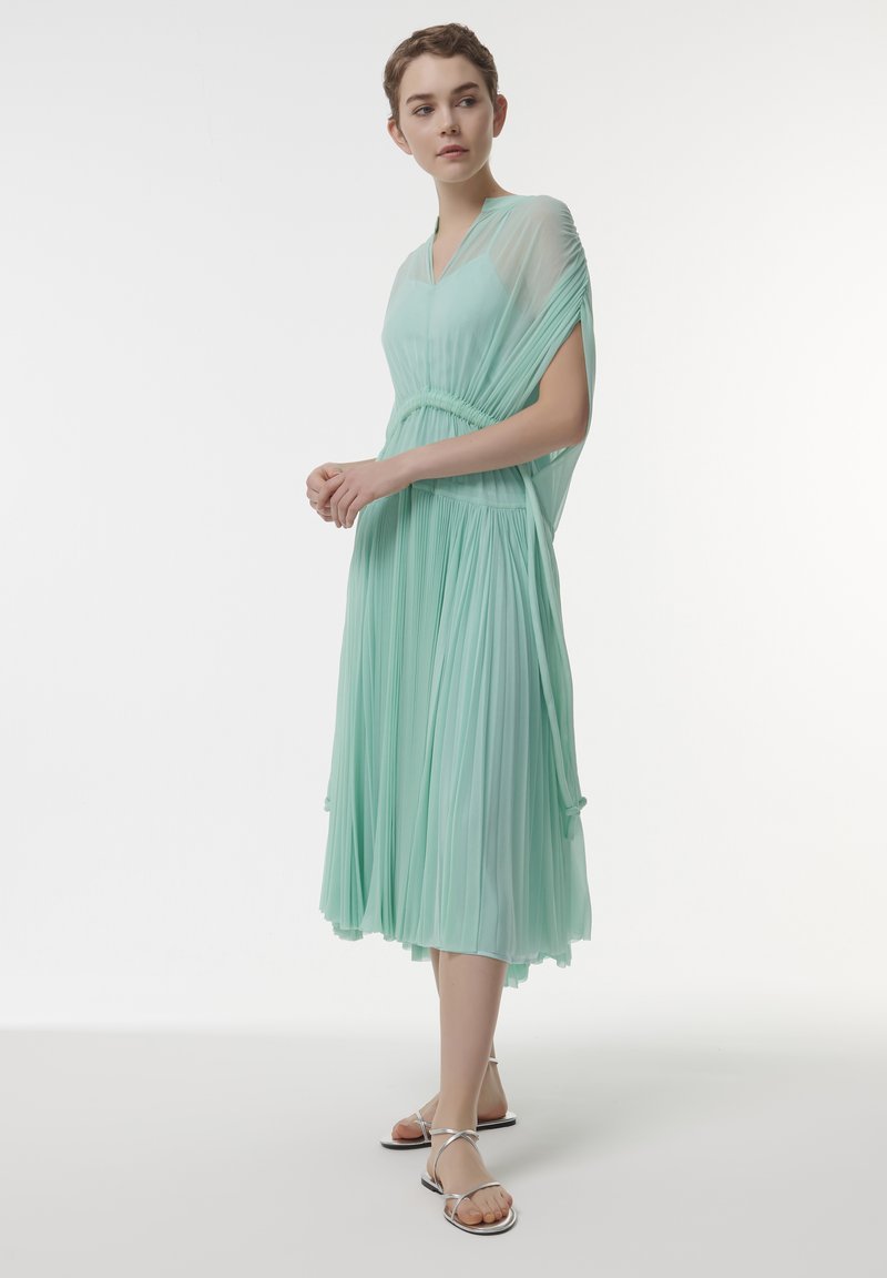 Vestido plisado verde menta con escote en V profundo, cintura ceñida y mangas fluidas. Combinado con sandalias plateadas de tiras. Tela suave y ligera.