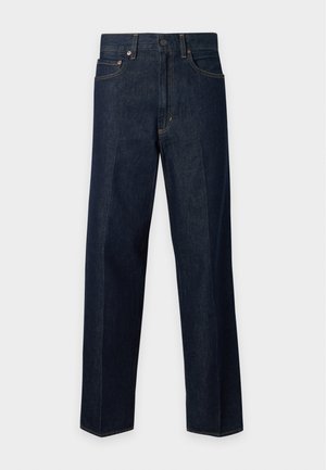Kvinde med langt blondt hår står iført en mørkegrå t-shirt, mørkeblå jeans og brune ankelstøvler mod en ensfarvet lys baggrund.