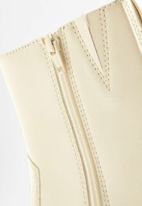 Gros plan sur une botte en cuir beige présentant une fermeture éclair verticale avec des détails cousus le long de la ligne de la fermeture et du bord supérieur.