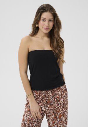 Jeune femme aux longs cheveux ondulés portant un haut noir sans bretelles, un pantalon à motif floral marron et blanc, des boucles d'oreilles en or et un bracelet.