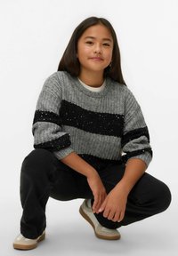 Szary sweter dziany z czarnymi cekinami w paski, noszony z czarnymi dżinsami i beżowymi butami. Modelka kuca i się uśmiecha.