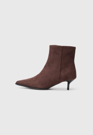 Bota de tornozelo em camurça castanha com bico apontado e um salto baixo e esculpido em preto. Costuras ao longo dos lados adicionam textura.