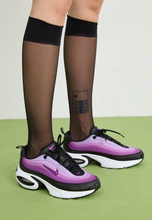 Ben iført sorte gennemsigtige knæstrømper og lilla-sorte hvide Nike-sneakers på grøn gulv med en tatovering på højre læg.