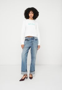 Sweatshirt branco cropped com logótipo turquesa, combinado com jeans de perna larga azul clara dobrados na bainha e saltos vermelhos escuros de bico fino.