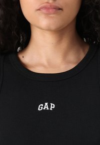 Crni rebrasti top bez rukava s okruglim izrezom i bijelim izvezenim logom "GAP" na prednjem dijelu.