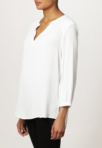 Vit blus med V-ringning, trekvartsärmar och texturerade axeldetaljer. Tillverkad av lättviktsmaterial med en avslappnad passform.