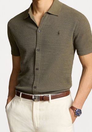 Man draagt een kort mouwenshirt in olijfgroen met een gestructureerd patroon, beige broek met een bruine leren riem en een bruine horloge met een blauw wijzerplaat.