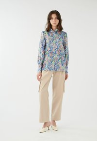 Blusa blu con motivo floreale, maniche lunghe e colletto a stand, abbinata a pantaloni cargo beige e mule col tacco color avorio.