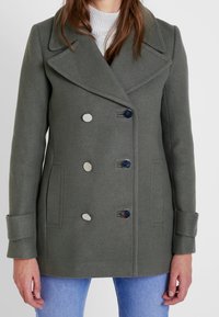 Abrigo de lana verde de doble botonadura con grandes solapas, botones plateados y bolsillos laterales. Presenta una tela texturizada y un ajuste entallado.