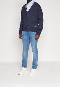 Mężczyzna w granatowym kardiganie na jasnoniebieskiej koszuli, w jasnoniebieskich dżinsach i białych trampkach stoi na tle białej ściany.