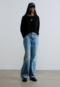 Μαύρο πουλόβερ με μακριά μανίκια, ανοιχτό μπλε τζιν παντελόνι με ευρεία ποδόγυρα και φθορές, και μαύρα παπούτσια τύπου slip-on. Συμπληρώνεται με μαύρο καπέλο με λογότυπο.