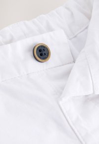 Tela blanca con superficie texturizada, que presenta un botón negro redondo con un borde beige y texto en relieve. El cuello tiene un detalle fruncido.