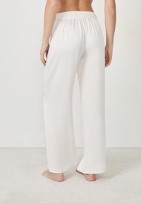Personne se tenant pieds nus sur un tapis, portant un pantalon en satin blanc ample et taille haute avec une ceinture élastique, vu de derrière.