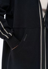 Czarna kurtka z zamkiem błyskawicznym, posiadająca fakturowany zamek i kremowe zdobienia wzdłuż rękawów i kieszeni. Gładki materiał z kieszeniami.