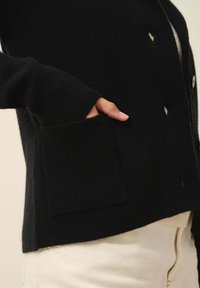 Czarny dzianinowy kardigan z przednią kieszenią, okrągłym dekoltem i zapięciem na guziki. Posiada teksturowane wykończenie oraz widoczne szczegóły ściegów.