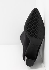 Chaussure noire à talon aiguille avec un bout pointu et une semelle en caoutchouc texturé arborant un motif floral. Le matériau semble être lisse et élastique.