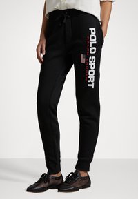 Černé tepláky mají šňůrku v pase, žebrované manžety a logo "POLO SPORT" v bílé a červené barvě s americkou vlajkou jako akcentem.