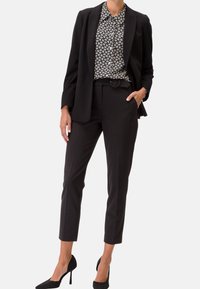 Blazer nero sartoriale sopra una camicia a righe bianco e nera, abbinato a pantaloni neri slim-fit e scarpe con tacco nere.