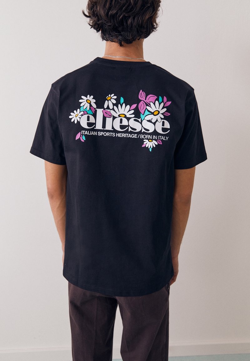 Personne portant un t-shirt noir avec un texte blanc "ellesse" et des motifs floraux colorés au dos, debout contre un mur clair uni.