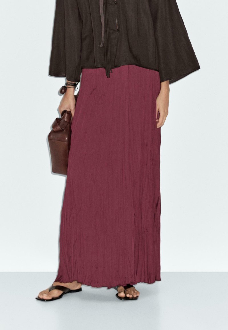 Jupe longue plissée bordeaux avec une finition texturée, associée à un haut ample foncé et des accessoires marron, portée avec des tongs noires.