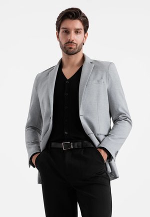 Blazer grigio chiaro con colletto a lancia, tasca singola, abbinato a una camicia nera con bottoni e pantaloni neri, indossato con una cintura.