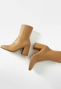 Botas de tobillo en color marrón claro con un acabado suave y brillante, punta afilada y tacón grueso. La costura es sutil y combina con el color de la bota.