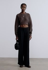 Pulover maro scurt, pantaloni negri cu picioare largi, ținând o geantă mică neagră. Design simplu, textură moale, culori neutre și croială ajustată.