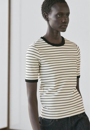 Giovane con indosso una maglietta a maniche corte a righe bianche e nere con rifiniture nere, in piedi contro uno sfondo grigio chiaro.