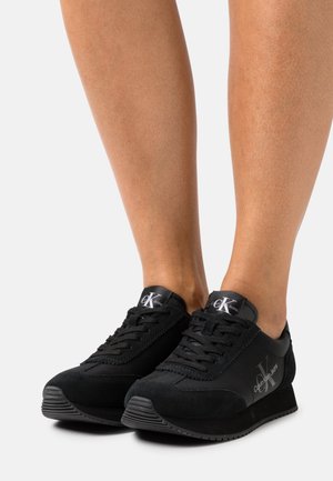 Persoon draagt zwarte Calvin Klein vetersneakers met verhoogde zool, staand tegen een effen lichte achtergrond.