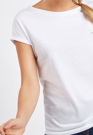 Kobieta ubrana w gładką białą koszulkę z krótkim rękawem z podwiniętymi mankietami oraz niebieskie dżinsy, przycięte na wysokości ramion i talii.