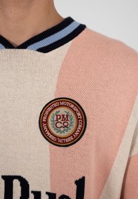 Sweter w pionowe kremowe i delikatnie różowe paski, z okrągłą bordowo-złotą naszywką z napisem "Prohibited Motorsport Company Berlin Germany" oraz czarno-niebieskim paskowanym kołnierzem.