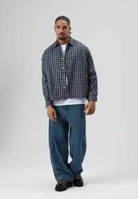 Kariertes, übergroßes Hemd in Marineblau und Schwarz, darunter ein weißes T-Shirt, kombiniert mit locker sitzenden blauen Jeans und schwarzen Plateau-Sneakern. Glatte Stofftextur.