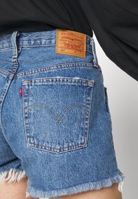 Levi's siniset farkkushortsit, joissa on revitty helma, erottuva takataskun ommel ja brändätty ruskea nahkapatch vyötäröllä.