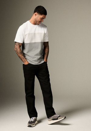 Homme aux bras tatoués portant une chemise blanche et grise à manches courtes, un pantalon noir et des baskets grises, regardant vers le bas les mains dans les poches.