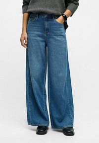 Persona que lleva unos vaqueros de denim azul holgados, zapatos oscuros y un suéter gris, con una mano en el bolsillo y una pulsera negra en la muñeca.